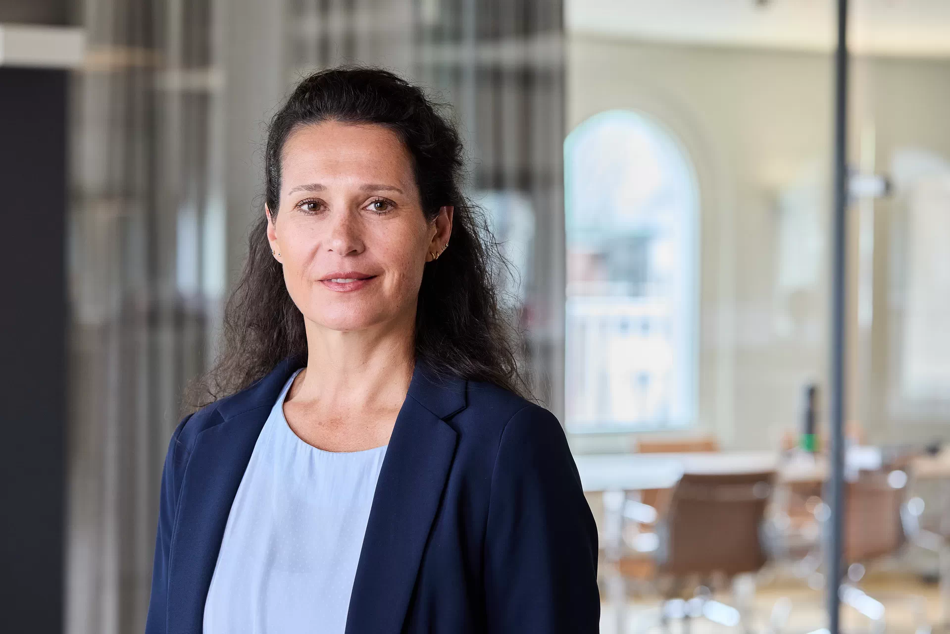 Dr Ulrike Handel steht vor einem verglasten B&uuml;ro, hat dunkelbraune Haare, tr&auml;gt einen blauen Blazer und schaut in die Kamera